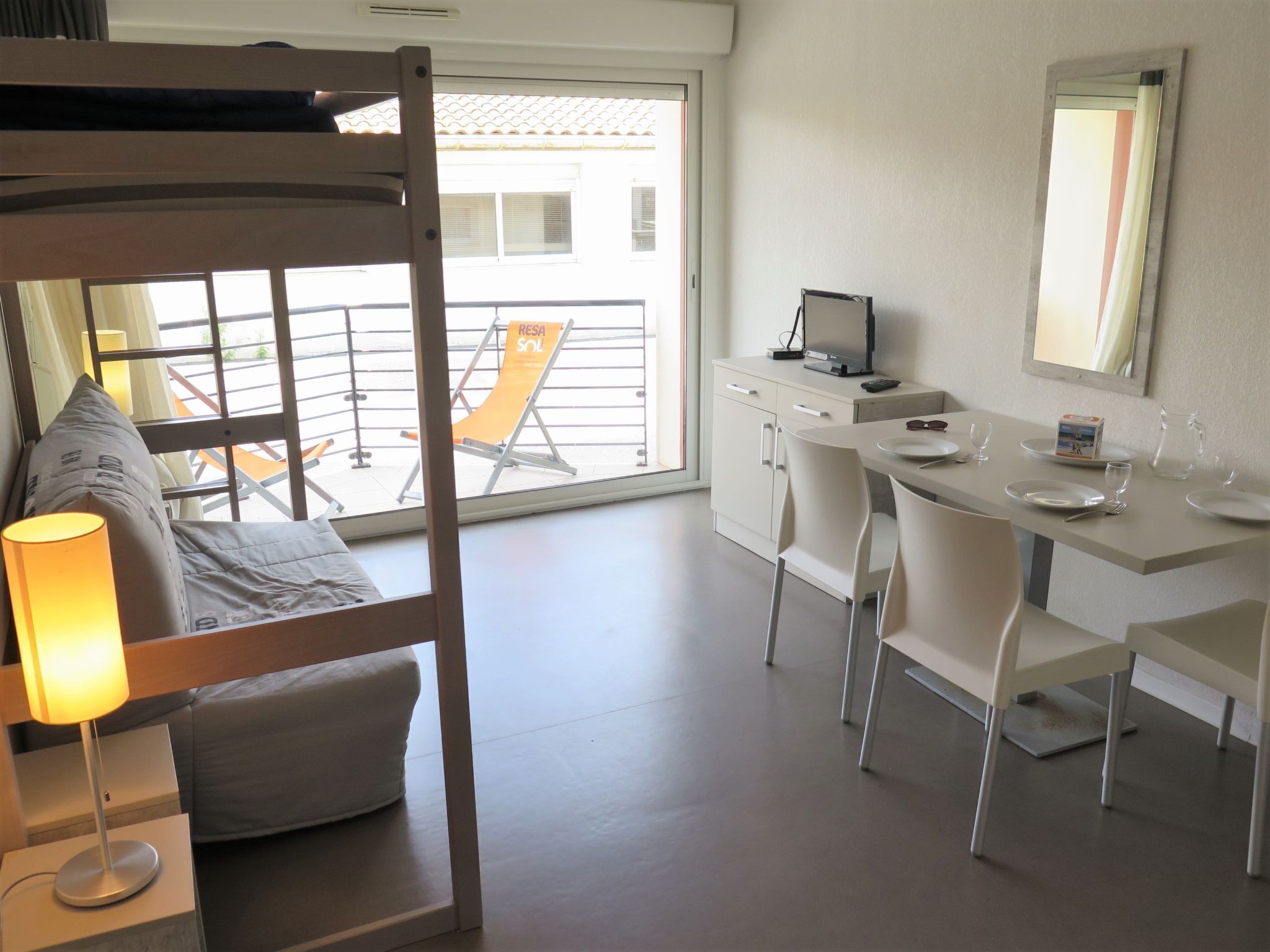 Quarto de hotel com cama beliche, escrivaninha com computador, mesa de jantar com cadeiras e varanda com cadeira e mesa.