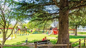 Ein Spielplatz mit verschiedenen Spielgeräten, einer Bank und einem großen Baum im Vordergrund.
