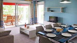 Uma área de refeições com mesa posta para seis, um sofá, uma televisão e uma varanda com cadeiras.