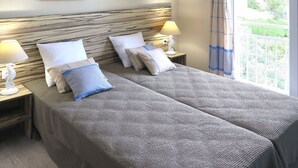 Quarto de hotel com duas camas, cabeceira de madeira, duas mesas de cabeceira com luminárias e dois quadros na parede retratando cenas de praia.