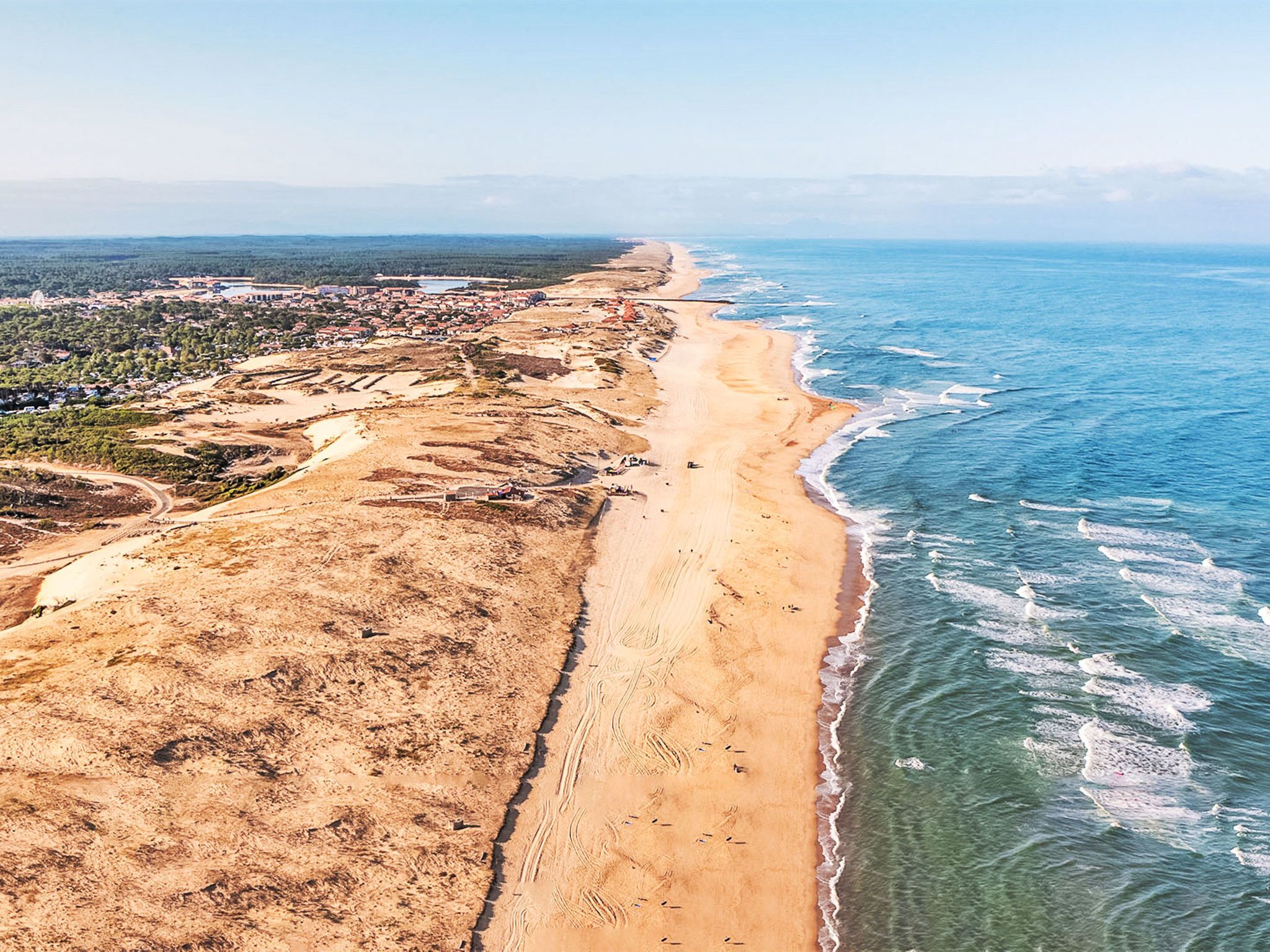 Oversigtsbillede af et kystområde med sandstrand, en lille bebyggelse og et klart, blå hav.