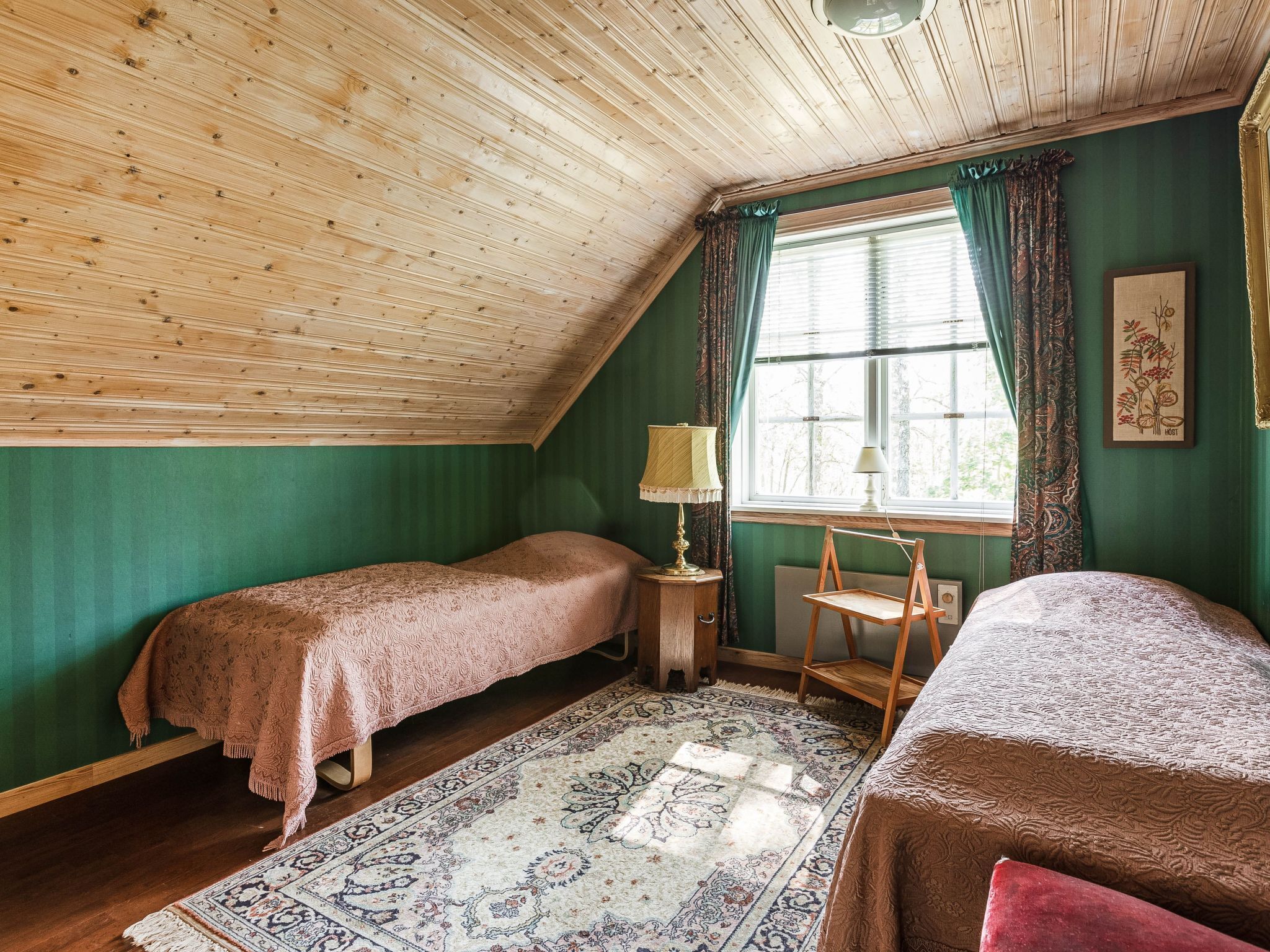 A cozy room with two beds, a wooden ceiling, a window with curtains, and a patterned rug.