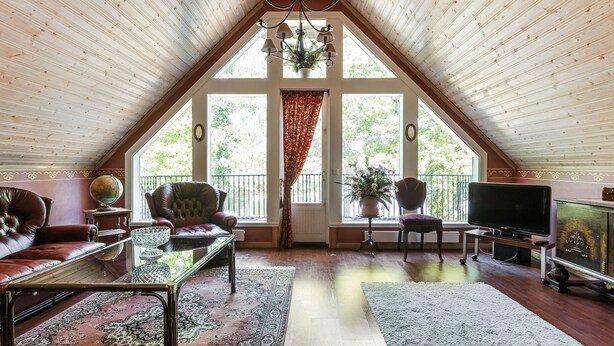 Ein geräumiges Dachzimmer mit geneigter Decke, großen Fenstern, einer Chandelieber, einem Ledersofa, einem Couchtisch, einem Fernseher und einem Schrank.