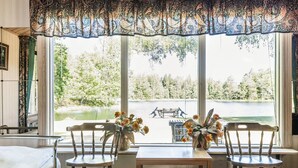Ein Zimmer mit einem Bett, zwei Holzstühlen, einem kleinen Tisch und Blick auf einen See und Bäume durch große Fenster.