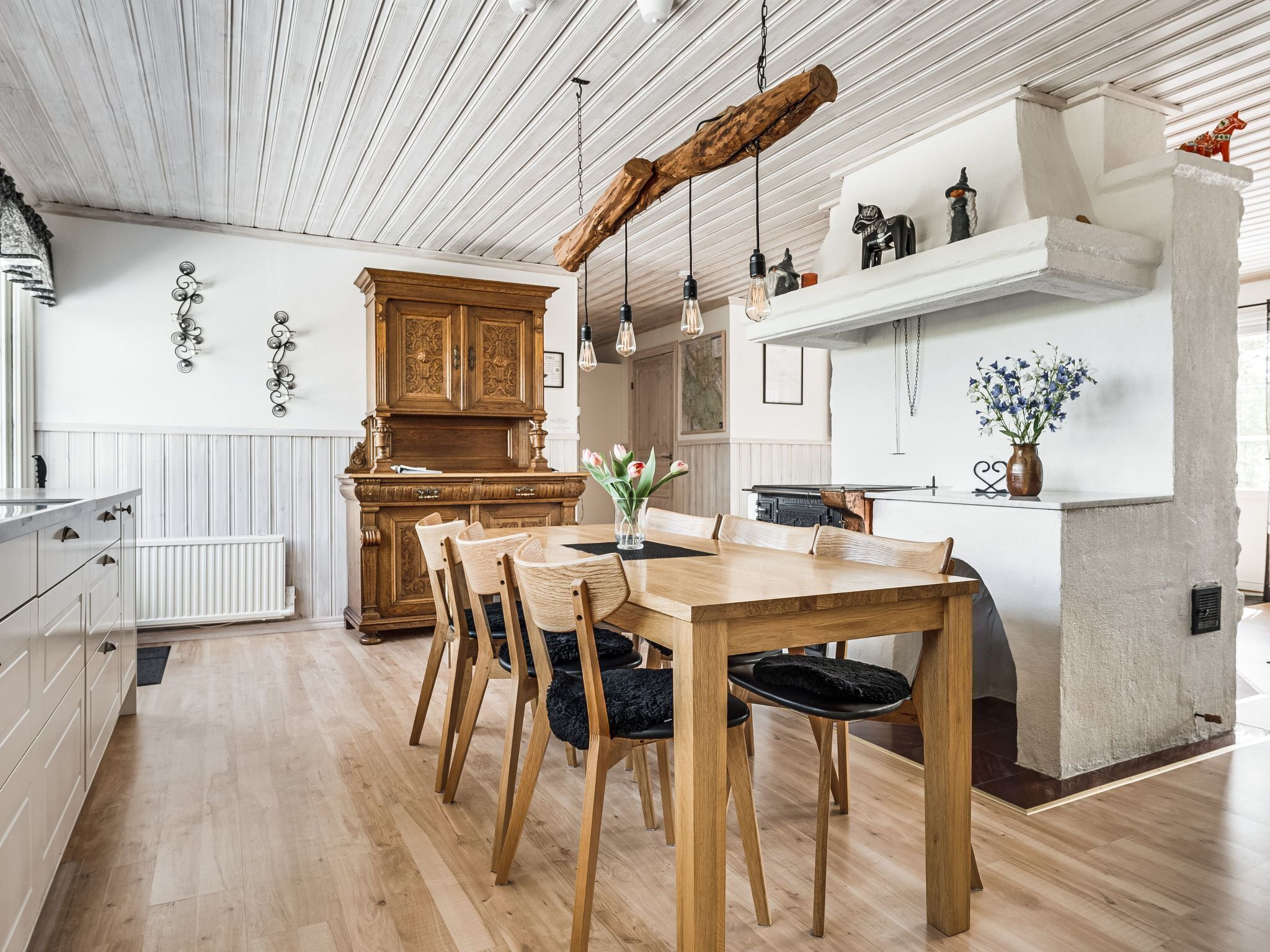A modern kitchen with wooden cabinetry, a dining table with chairs, pendant lights, and a white island with a vase of flowers.