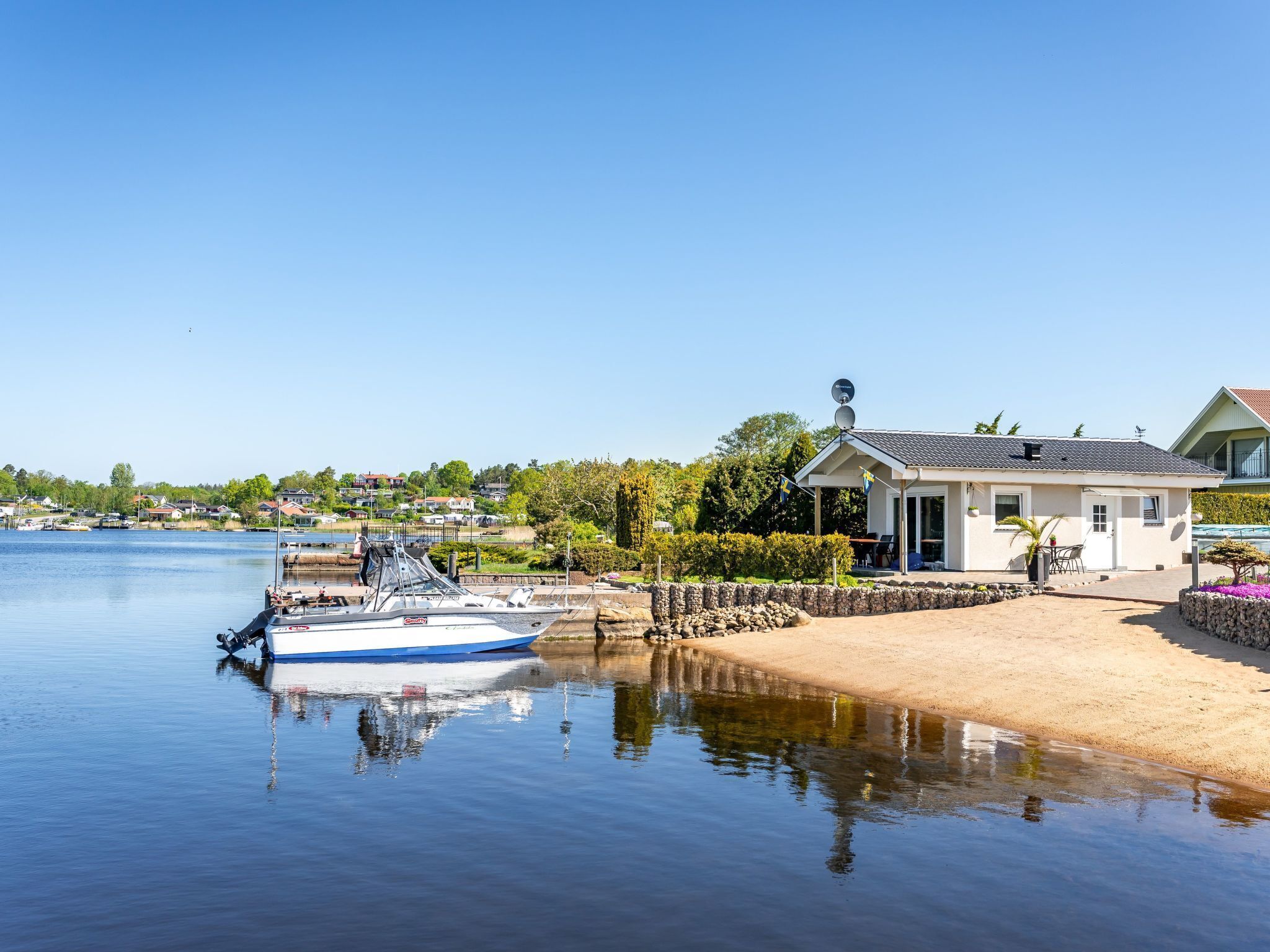A boat docked at a calm waterfront with a house featuring a satellite dish and a small garden.