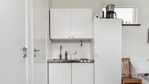 Une cuisine de taille réduite, avec des meubles blancs, un évier et une cuisinière. La pièce adjacente contient une chaise en bois et un lit avec un oreiller blanc.