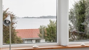 Une vue à travers une fenêtre, avec une lampe blanche posée sur le rebord, donnant sur une étendue d’eau et un bâtiment dont le toit est recouvert de tuiles rouges.