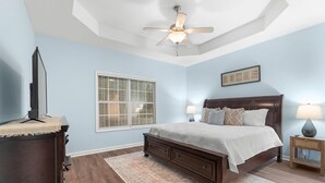 Un dormitorio con cama, ventilador de techo, ventana con persianas, mesita de noche con lámpara y cómoda.