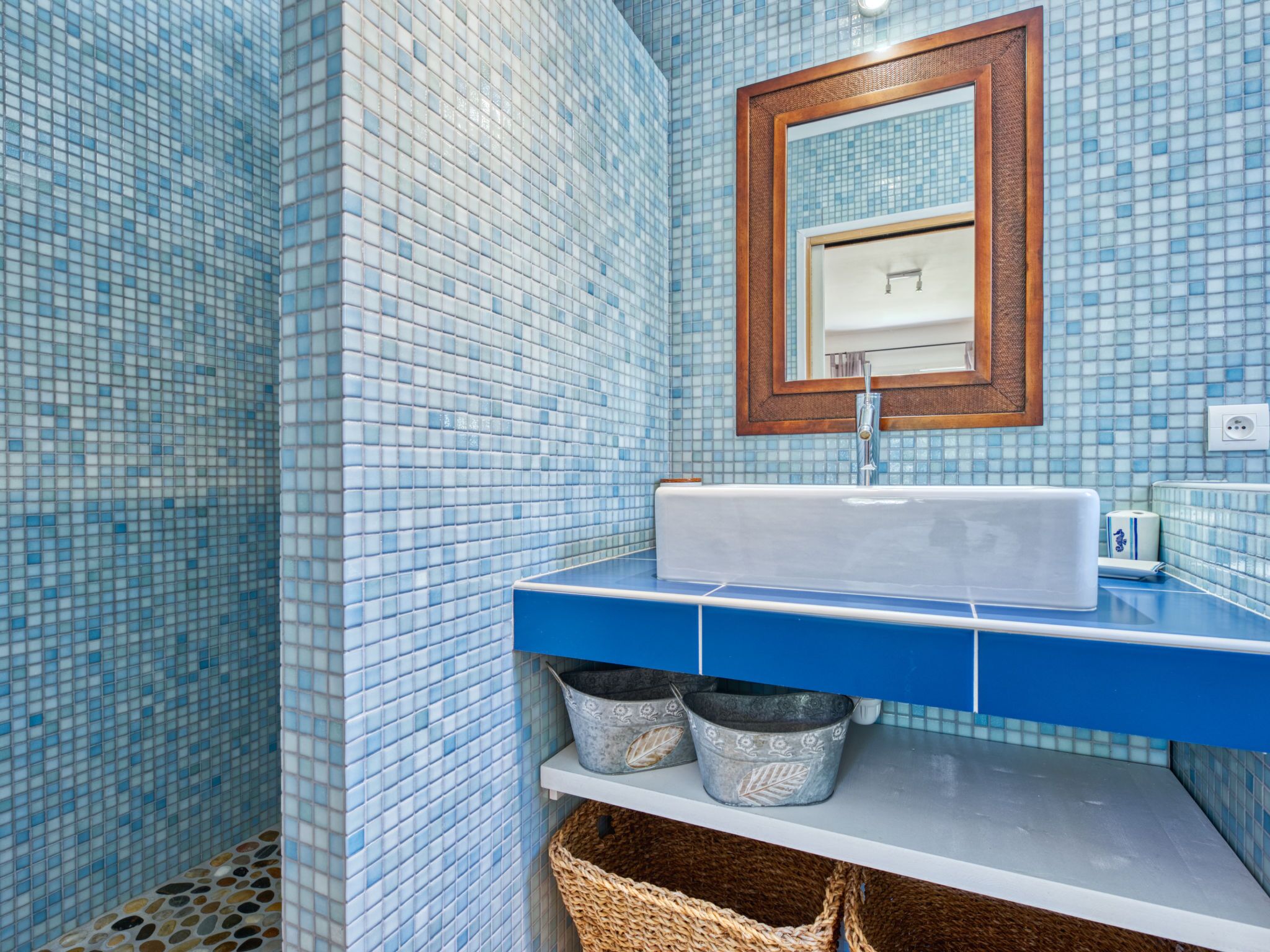 A bathroom with a blue tiled wall, a white sink, a wooden-framed mirror, and a shelf with two metal buckets.