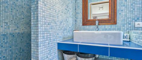 A bathroom with a blue tiled wall, a white sink, a wooden-framed mirror, and a shelf with two metal buckets.