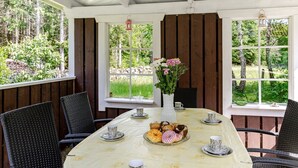 Ett takförsett utomhusområde med ett bord dukat för frukost, omgivet av stolar, och utsikt över ett skogsområde.