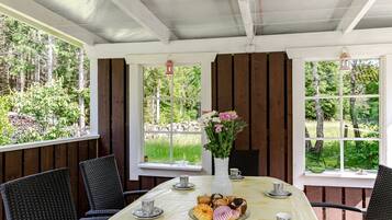 Un espace extérieur abrité, aménagé pour prendre les repas, avec une table dressée pour le petit-déjeuner, entourée de chaises, et offrant une vue sur une zone boisée.