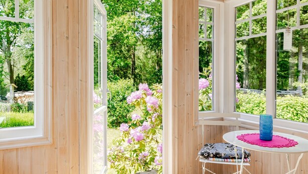 Ett rum med stora fönster, ett runt bord med en blå vas och en stol med ett blommigt kuddöverdrag.