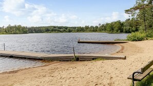 En sandstrand med en träbrygga som sträcker sig ut i en sjö, omgiven av träd.
