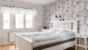 Une chambre avec un lit blanc, une table de chevet, une commode, une fenêtre avec un rideau et un mur orné d’un motif en forme d’étoiles.