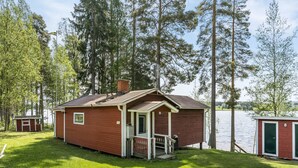 Une petite cabane dotée d’une véranda, d’une cheminée et offrant une vue sur un lac entouré d’arbres.