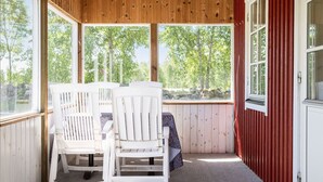A covered outdoor seating area with white chairs and a table, large windows offering a view of trees and a stone wall. - Skärvudde Huset by Interhome (Kalvsvik)