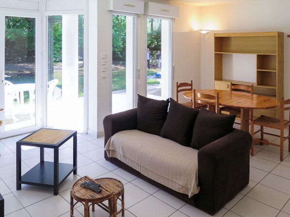 A living room with a sofa, a small table, a dining table with chairs, and a large window with a view of greenery.