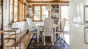 A dining area with a wooden ceiling, large windows, a white table with chairs, a bookshelf, and a radiator.