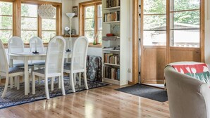 A cozy room with a wooden ceiling, a dining table with chairs, a bookshelf, and a large window.