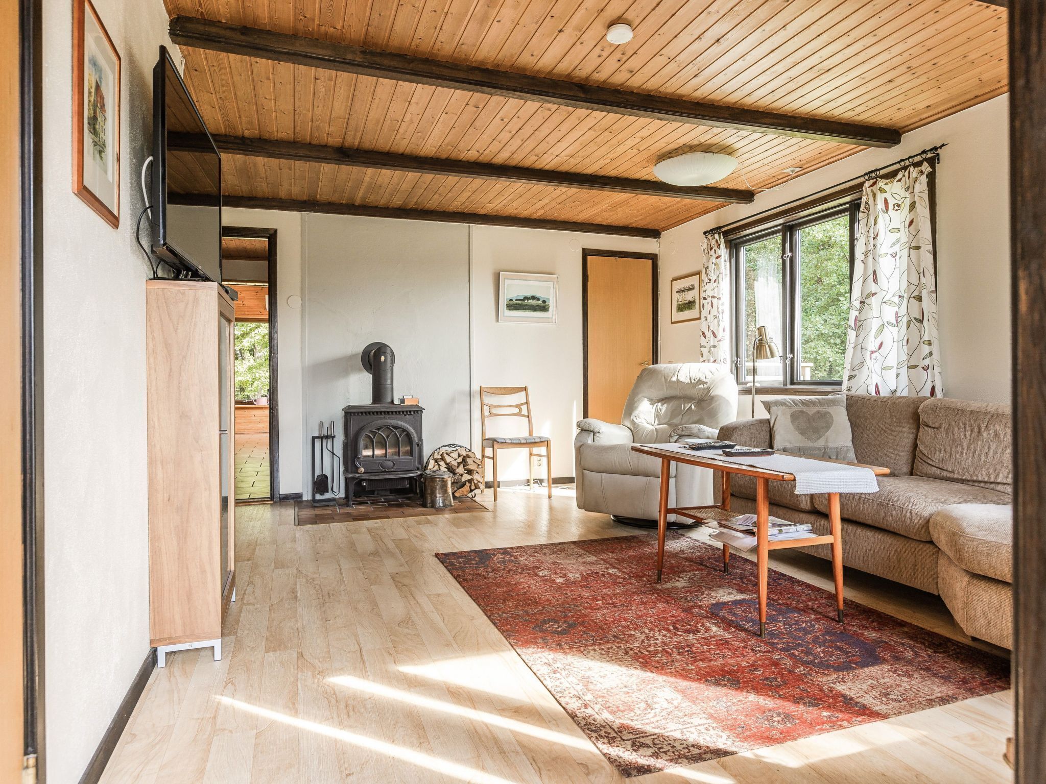 A cozy living room with a wooden ceiling, a fireplace, a sofa, and a large window with curtains.