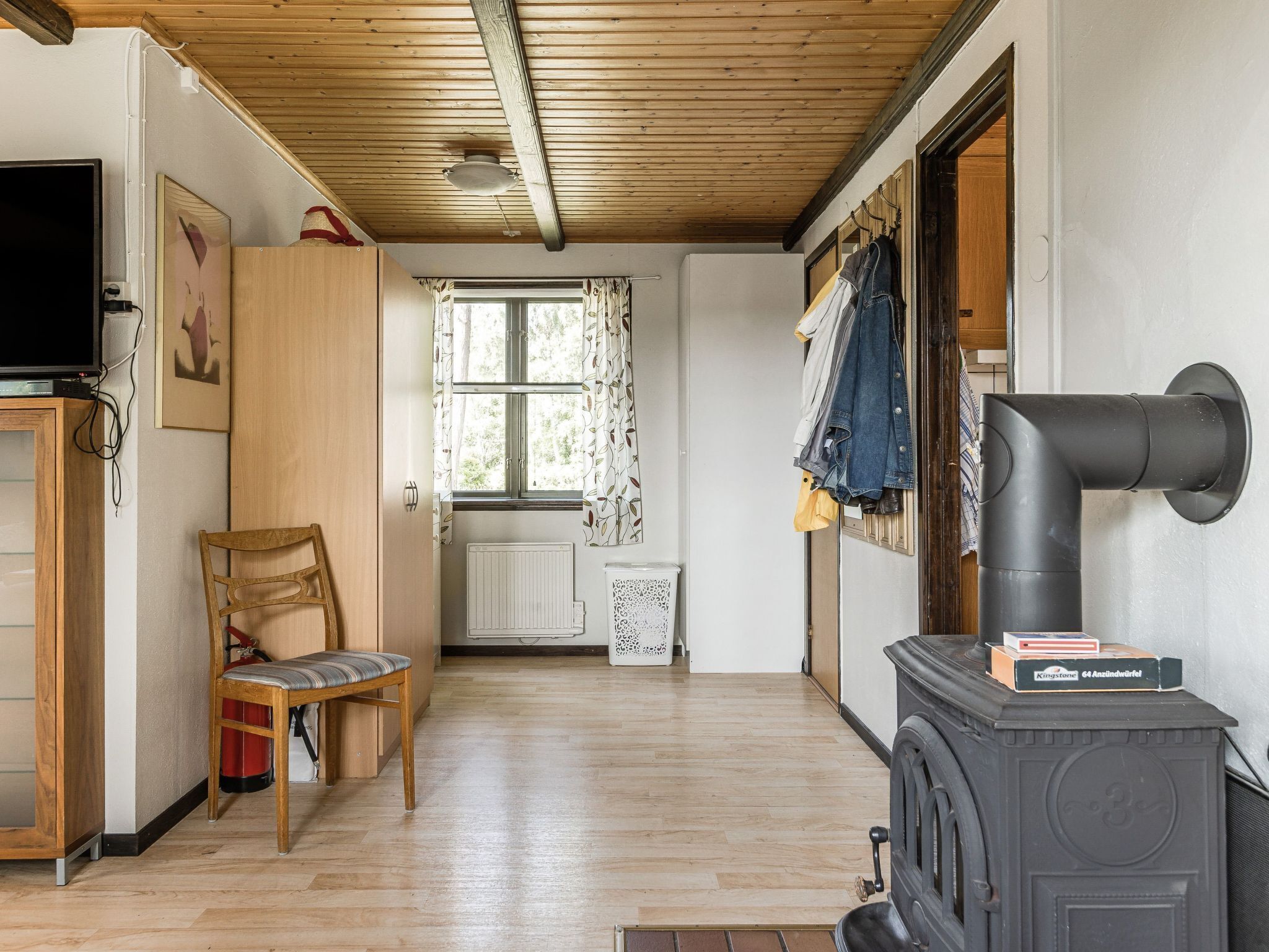 Una habitació acollidora amb sostre de fusta, un televisor, una cadira, una finestra amb cortines, un radiador i una estufa negra.