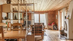 A cozy cabin interior with wooden floors, a dining area, a seating area with a fireplace, and a large window offering a view of the outdoors.
