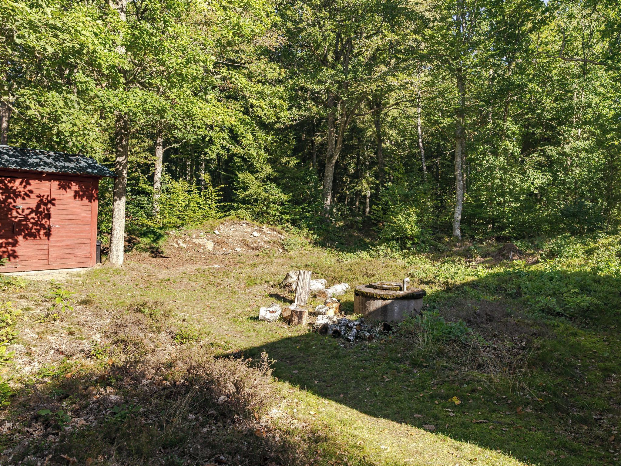 A red wooden cabin in a forested area with a pile of logs and a concrete structure nearby.