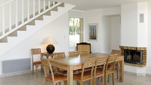 A dining room with a wooden table and chairs, a lamp, a fireplace, and a staircase.