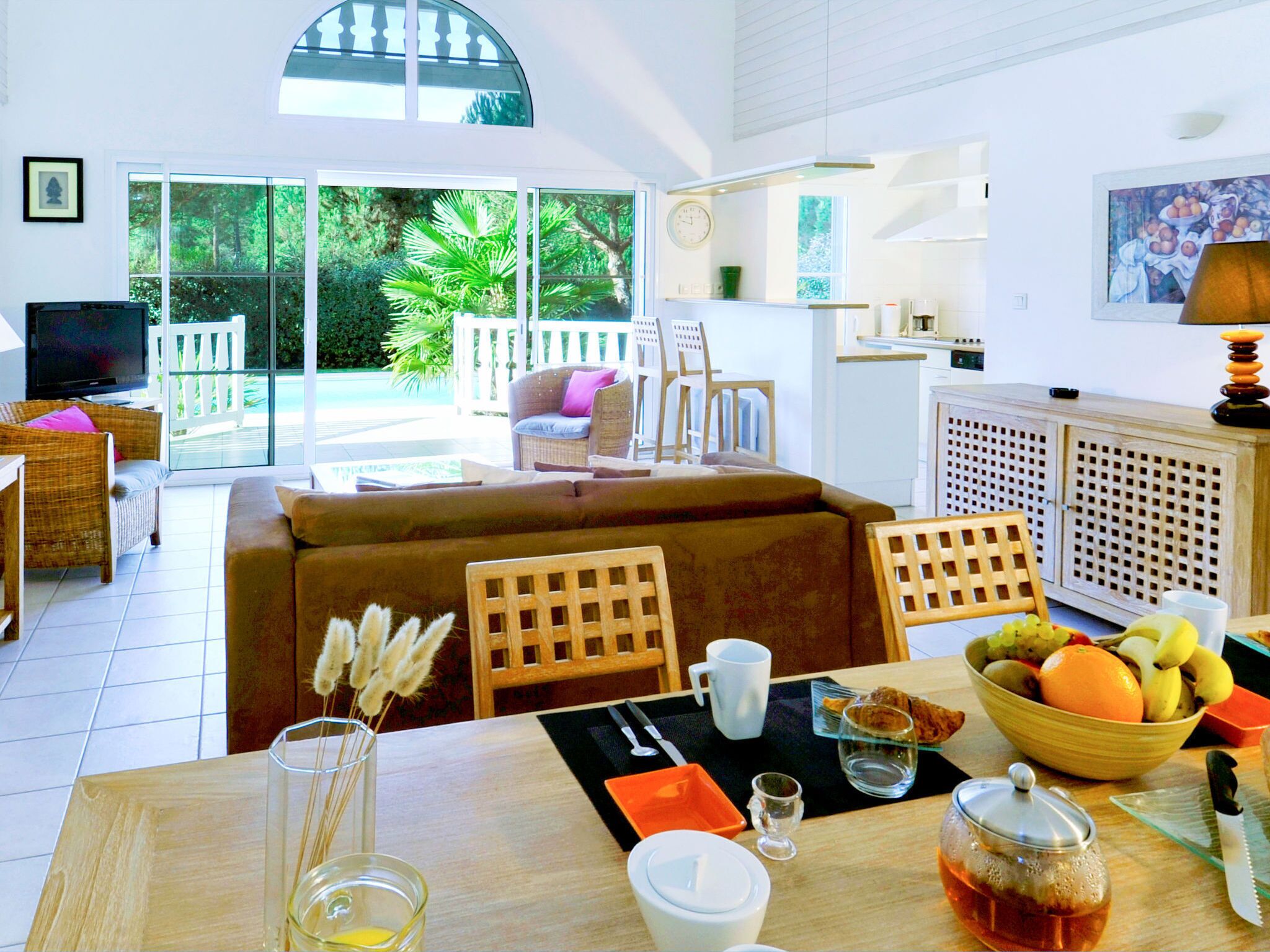 Un vaste espace de vie comprenant un canapé, des fauteuils, une table à manger avec un panier de fruits, et un téléviseur en arrière-plan.
