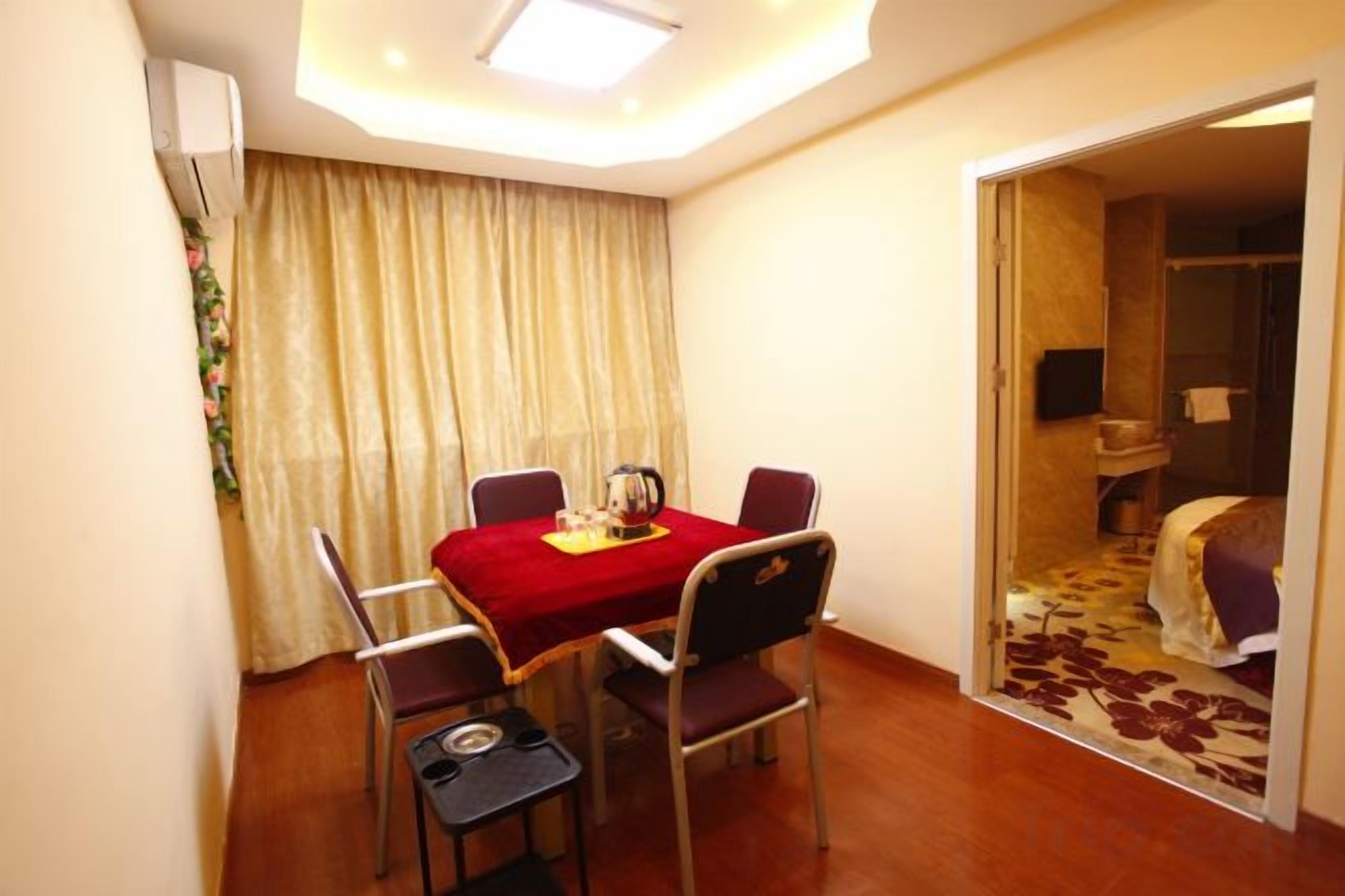 A hotel room with a dining table, four chairs, a small side table, a bed with a floral bedspread, and a bathroom visible through an open door.