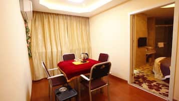 A hotel room with a dining table, four chairs, a small side table, a bed with a floral bedspread, and a bathroom visible through an open door.