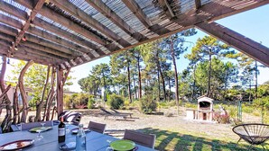Ein Essbereich im Freien mit einer Pergola, Tischen gedeckt mit Tellern und Weingläsern, und Blick auf eine bewaldete Landschaft mit einem Grill im Hintergrund.