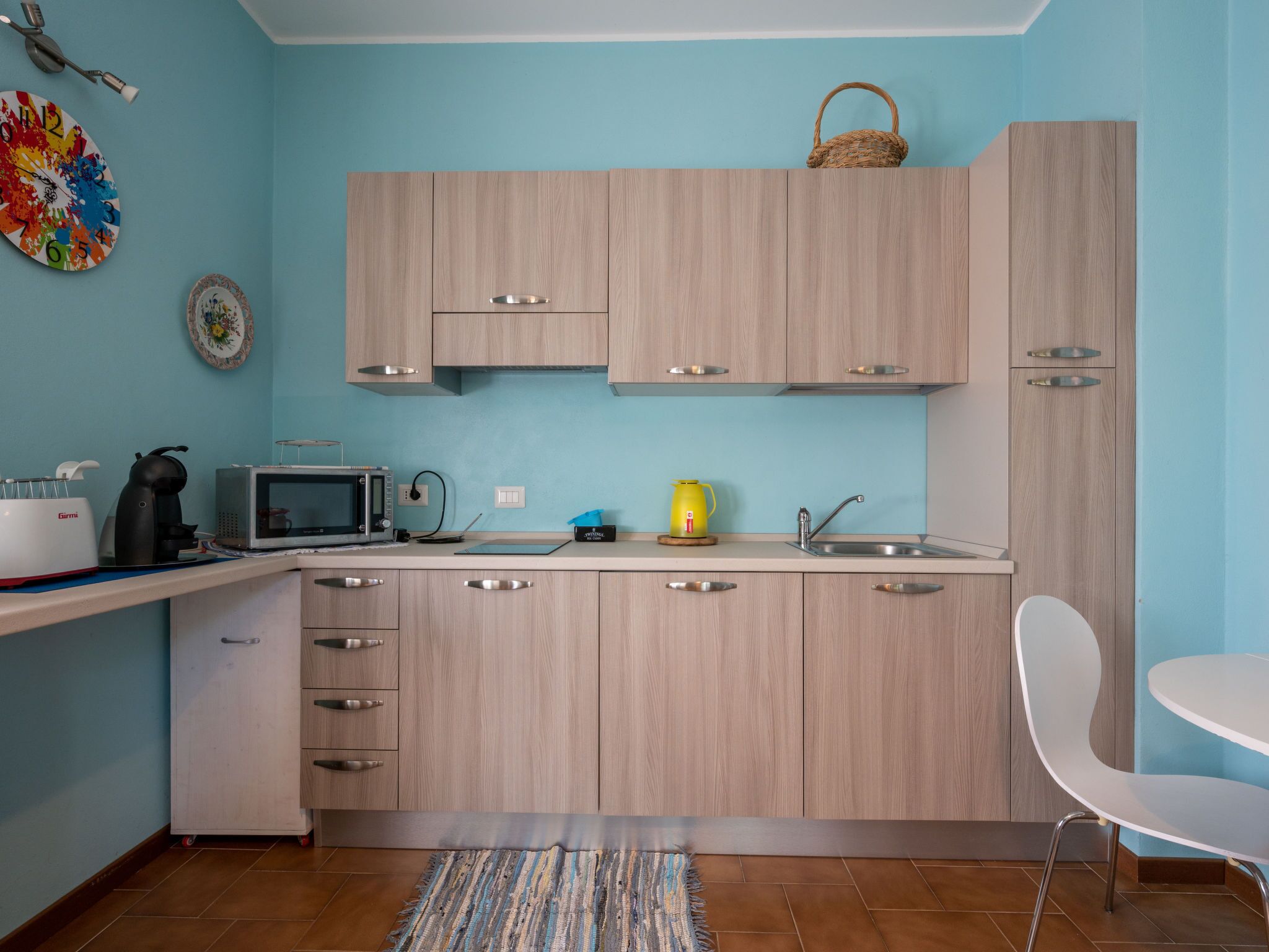 A modern kitchen with light wood cabinets, a microwave, kettle, and a sink.