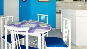 Uma área de refeições com uma mesa redonda posta para quatro pessoas, cadeiras brancas com almofadas azuis, um quadro na parede e uma cozinha ao fundo.