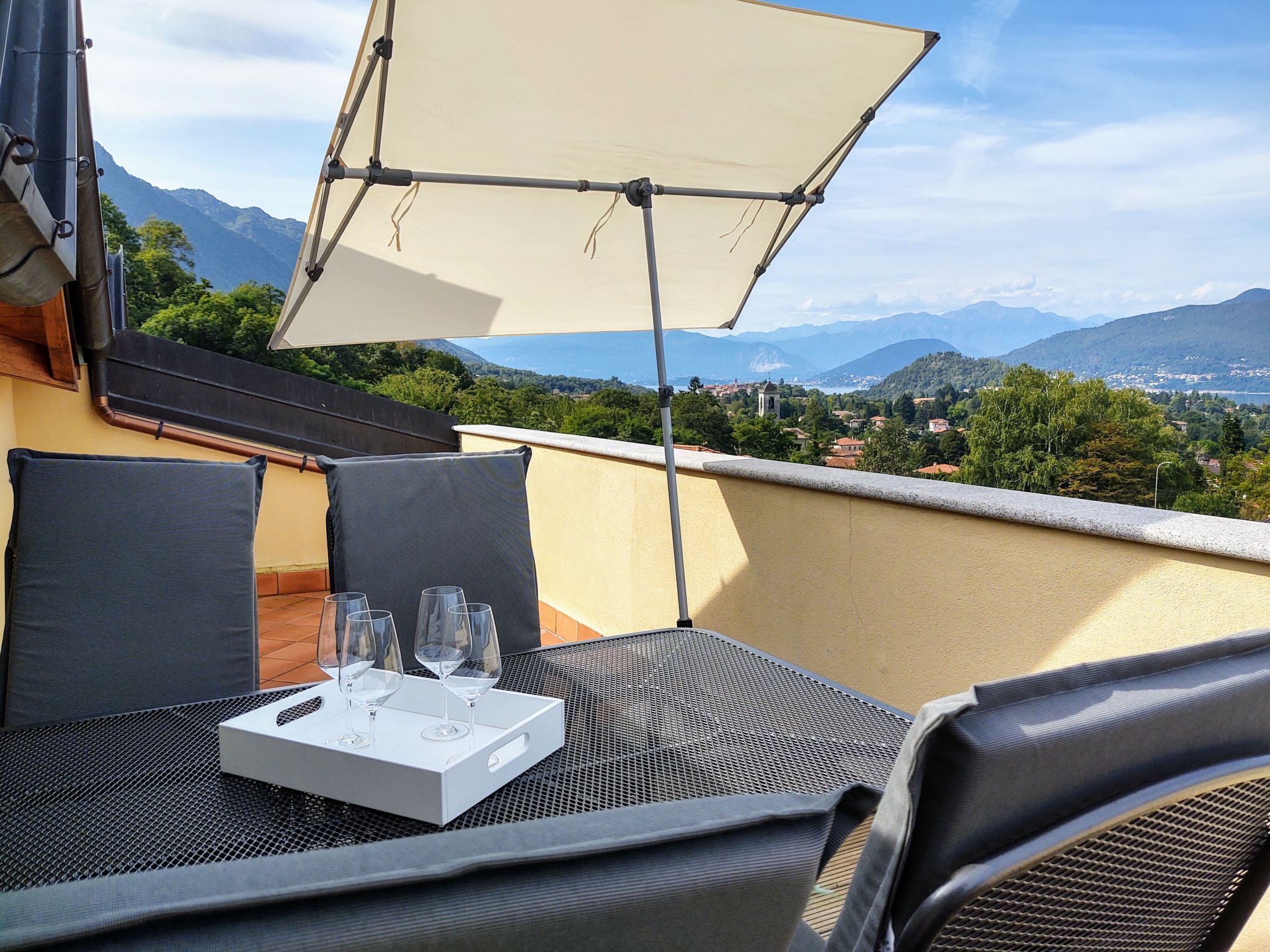Un balcón con una mesa lista para dos, una bandeja blanca con vasos y un toldo que da sombra.