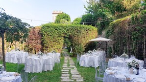 Un espacio para bodas al aire libre con varias mesas redondas dispuestas sobre un camino de piedra, rodeado de follaje y un muro de piedra.