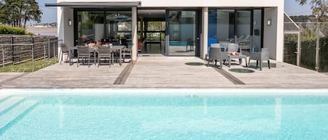 A modern house with a swimming pool, a patio with outdoor furniture, and a glass door leading to the pool area.