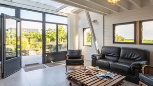 Un salon moderne doté d’une grande porte vitrée donnant sur un espace extérieur, d’un canapé en cuir noir et d’une table basse en bois sur laquelle reposent un magazine et un croissant.