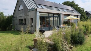 Une maison moderne dotée d’un toit métallique incliné, de grandes fenêtres et d’un espace patio aménagé avec du mobilier d’extérieur et un barbecue.