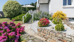Ein modernes Haus mit einer Steintreppe, die zu einer weißen Tür führt, umgeben von bunten Blumenbeeten und einem großen Baum.