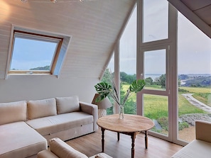 Ein Zimmer mit schrägen Wänden, einer Couch, einem Holztisch und großen Fenstern mit Blick auf eine Landschaft.