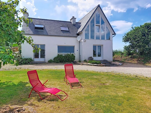 Une maison moderne à deux étages, dotée d’un toit en pente, de grandes fenêtres et d’une pelouse bien entretenue, agrémentée de deux chaises rouges.