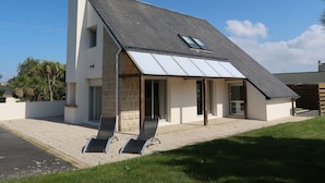 Een modern herenhuis met twee verdiepingen, een schuin dak, een groot raam en een terras met twee loungeschotels.
