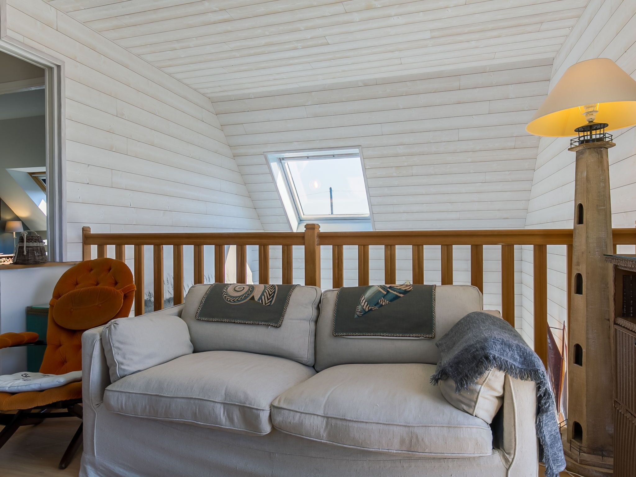 A cozy loft bedroom with a wooden ceiling, a sofa with decorative pillows, a lamp, and a view through a skylight.