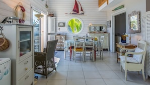 A cozy, well-lit room with a dining area, a wicker chair, a white cabinet, and a round window offering a view of greenery. - Penn an Aod by Interhome (Kerlouan)