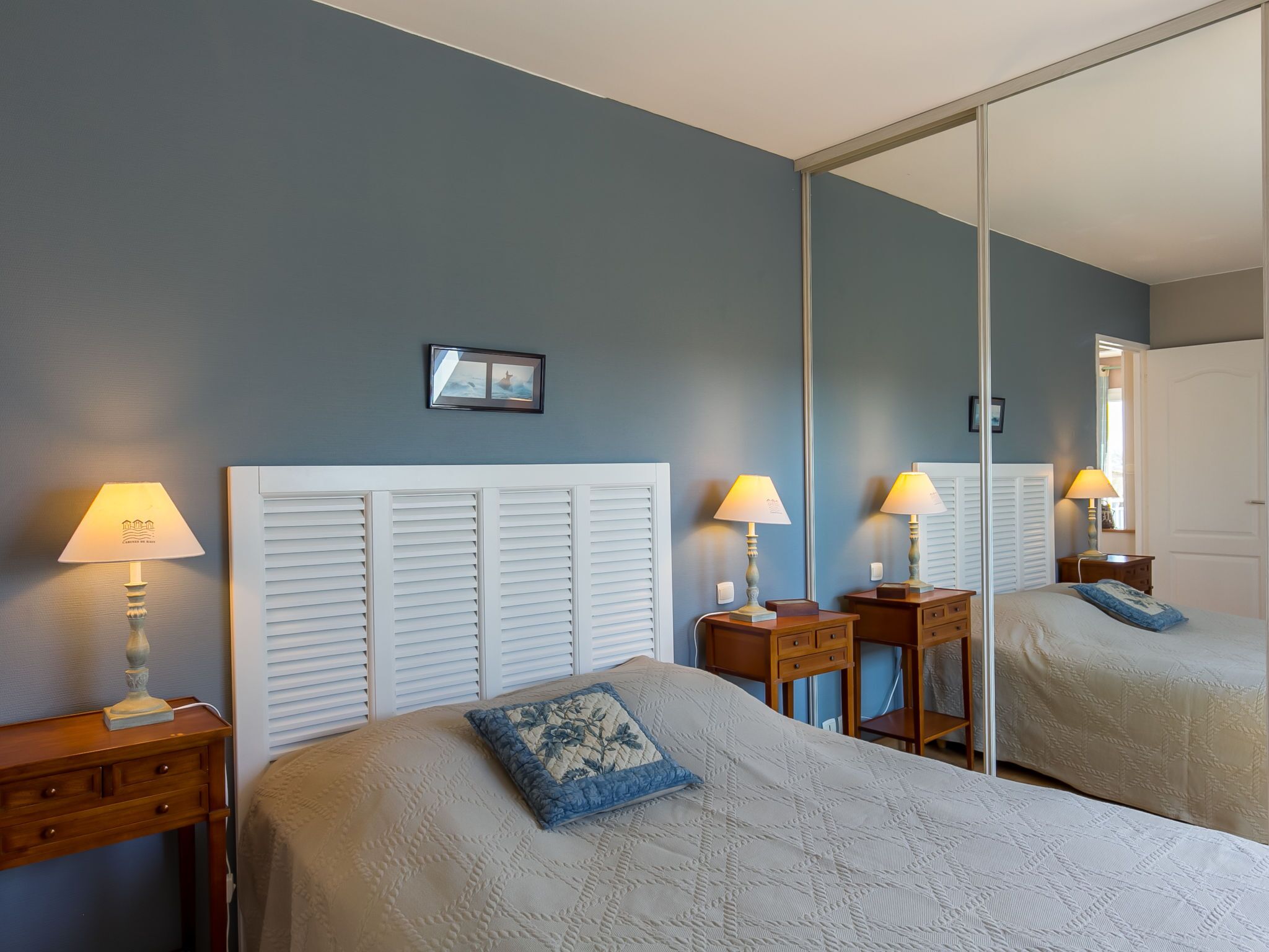 Un dormitorio con un espejo grande, dos mesitas de noche con lámparas, una cama con almohada estampada y un cuadro en la pared.