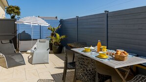 An outdoor patio with a table set for breakfast, including yellow bowls, a bottle of orange juice, and a basket of pastries. There are two chairs and a striped umbrella providing shade. - Penn an Aod by Interhome (Kerlouan)