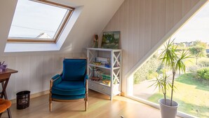 Une pièce chaleureuse avec une lucarne, un fauteuil bleu, une bibliothèque et une vue sur la verdure à l’extérieur.